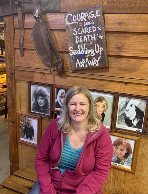 author at restaurant in front of sign saying "Courage is being scared to death and saddling up anyway."